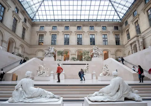 Louvre Museum Interior gallery of the Louvre Museum with classical marble sculptures and a grand staircase under a glass ceiling.