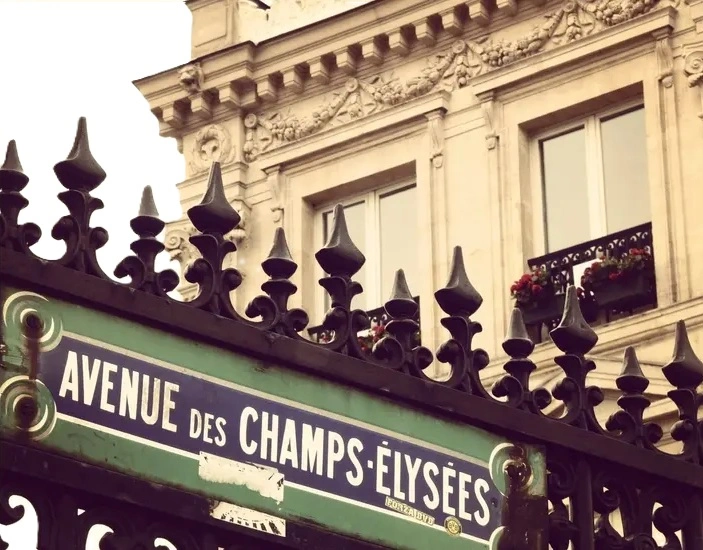 Champs-Elysees “Avenue des Champs-Élysées” mounted on an ornate iron fence, with a classic Parisian building featuring carved stone façades and flowered balconies in the background.